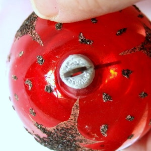 Op de afbeelding: Rode glazen kerstbal met zwarte glitteraccenten en een zilveren metalen hanger. De hanger heeft de tekst "West Germany" erop.