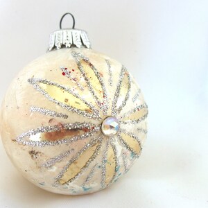 May include: Cream-colored Christmas ornament with a silver glitter design and a faceted jewel center. The ornament has a silver metal cap and a loop for hanging. The design features gold and silver accents.