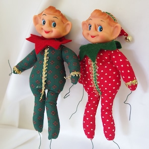 Puede incluir: Dos adornos de elfo de Navidad vintage con colgadores de alambre. Un elfo lleva un traje estampado en verde y rojo con ribete dorado y un cuello rojo. El otro elfo lleva un traje estampado en rojo y blanco con un cuello verde.