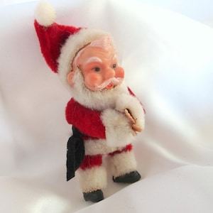May include: A vintage Santa Claus figurine made of red felt and white fur. He is wearing a red and white suit with a black belt and a white beard. He is holding a small brown object in his hand.