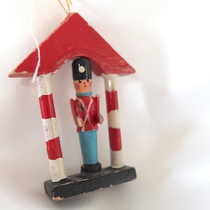 Puede incluir: Un adorno navideño de madera con forma de bastón de caramelo rojo y blanco con un pequeño soldado de madera de pie en el medio. El soldado lleva un abrigo rojo, pantalones azules y un sombrero negro.