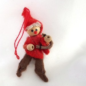 May include: A red felt Christmas ornament depicting a gnome holding a wooden stick with wooden beads. The gnome has a wooden head with a painted face and a red nose.