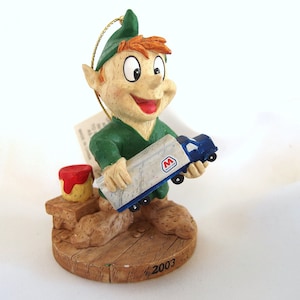 Puede incluir: Una figurita de elfo verde sosteniendo un camión de juguete azul y blanco con la letra "M" en el lateral. El elfo sonríe y tiene un bote de pintura rojo y amarillo a su lado. La base de la figurita es marrón y tiene el año "2003" inscrito en ella.