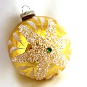 May include: A gold Christmas ornament with a white snowflake design and a green jewel in the center.