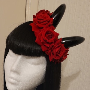 May include: A black headband with two horns and red roses. The headband is worn on a mannequin with black hair.