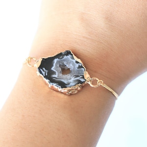 Puede incluir: Un brazalete de cadena dorada con un colgante de ágata negro y blanco. La rebanada de ágata tiene una forma natural irregular y un centro blanco.