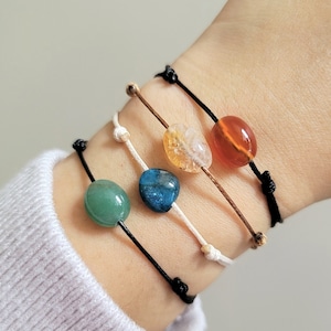 Könnte beinhalten: Vier Armbänder mit unterschiedlich farbigen Steinen am Handgelenk einer Person. Die Steine sind grün, blau, gelb und orange.