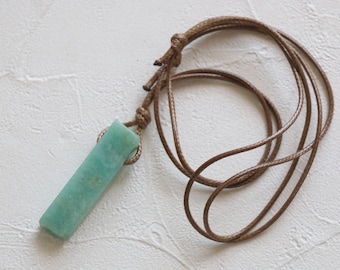 May include: A rectangular, light teal pendant necklace with a brown cord. The pendant is a solid, smooth stone. The cord is knotted at the top and hangs in a loop. The background is a textured white surface.