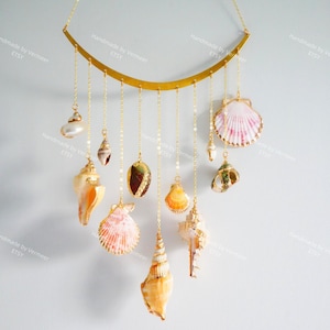 May include: A wall hanging with a gold-coloured metal bar, from which various seashells are suspended by gold chains. The seashells are in shades of white, pink, orange, and brown. The background is a light blue wall. The seashells are of various sizes and shapes.