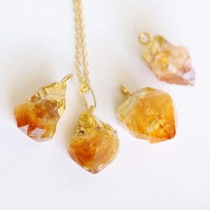 May include: Four gold-plated citrine crystal pendants, three of which are hanging from a gold chain. The crystals are all different sizes and shapes, and they have a warm, golden color.