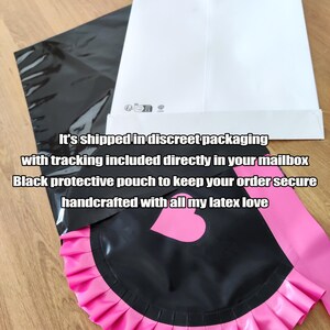 May include: Black latex apron with a pink ruffled trim and a heart-shaped detail. The apron is paired with a pink sash. The image also shows a black protective pouch and a white envelope.