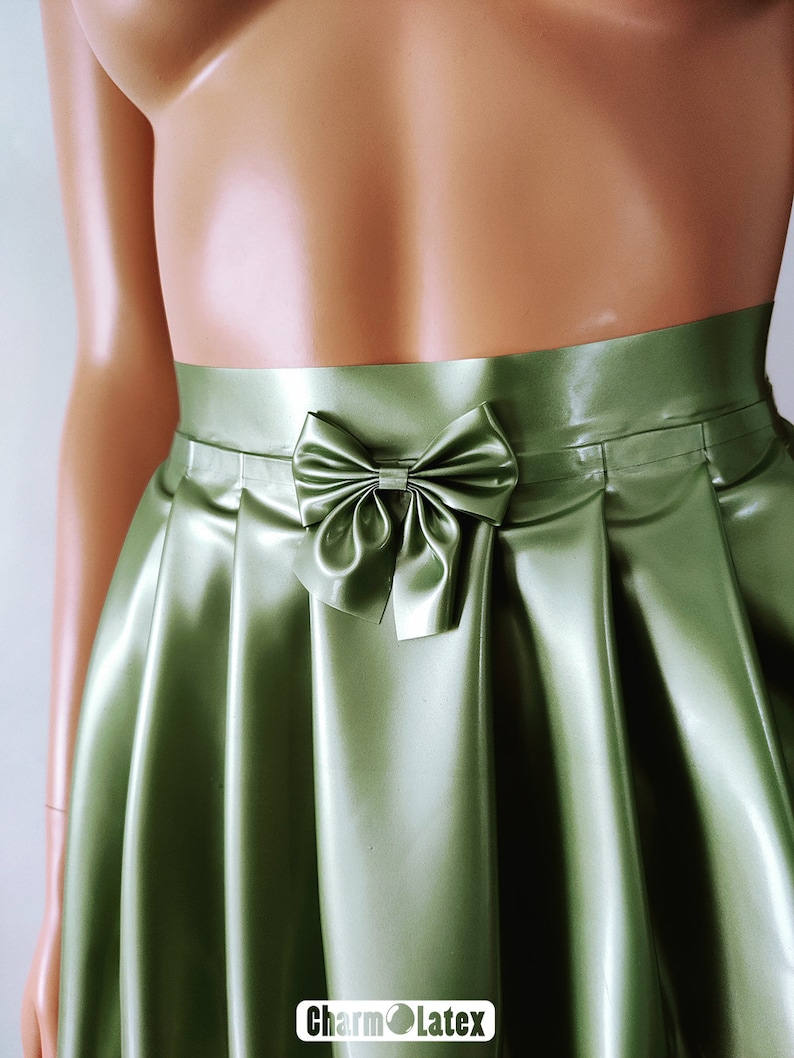 May include: A green satin skirt with a bow detail at the waist. The skirt has a pleated design and a shiny finish. The logo "Charm Latex" is visible at the bottom right.