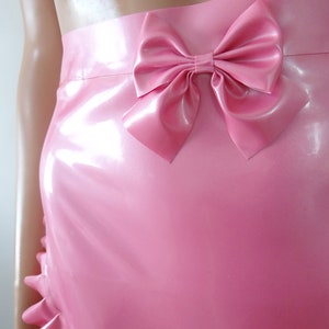 May include: A pink latex skirt with a large bow detail at the waist. The skirt has a ruffled hemline and a glossy finish. The image shows a close-up of the skirt, highlighting its smooth texture and design.
