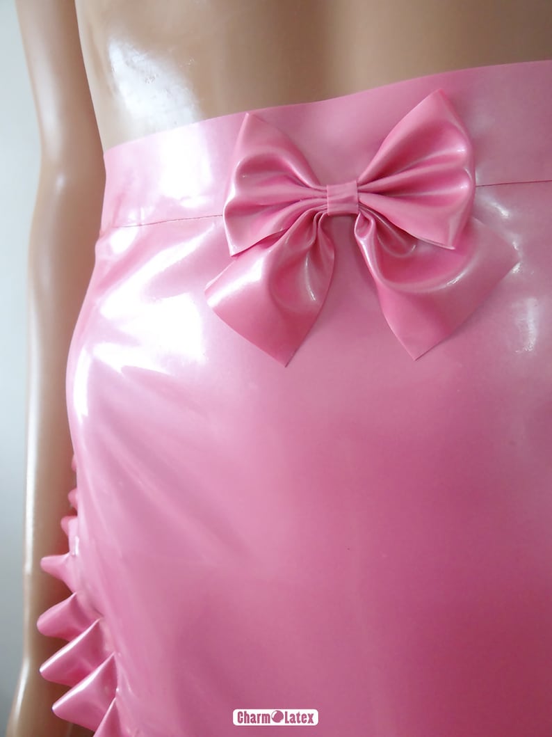 May include: A pink latex skirt with a large bow detail at the waist. The skirt has a ruffled hemline and a glossy finish. The image shows a close-up of the skirt, highlighting its smooth texture and design.