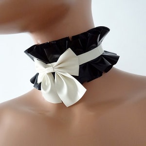May include: Black and white ruffled choker with a cream-colored bow. The choker features a wide, ruffled collar and a contrasting cream-colored bow at the front. The brand name "Charm Latex" is visible at the bottom.