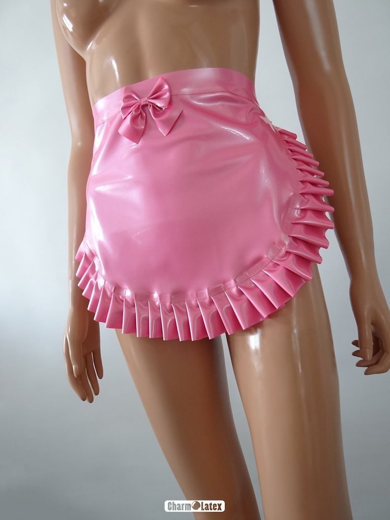 Peut inclure: Un tablier rose en latex avec un nœud à la taille et un ourlet à volants. Le tablier est brillant et semble être fait d'un matériau lisse. Le tablier est présenté sur un mannequin.