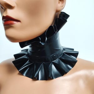 Könnte beinhalten: Schwarzer Latex-Rüschenkragen mit hohem Halsausschnitt. Der Kragen hat mehrere Lagen plissierter Rüschen und eine glatte, glänzende Oberfläche. Der Markenname "Charm Latex" ist unten sichtbar.