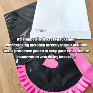 May include: Black latex apron with a pink ruffled trim and a heart-shaped detail. The apron is paired with a pink sash. The image also shows a black protective pouch and a white envelope.