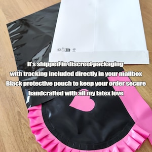 May include: Black latex apron with a pink ruffled trim and a heart-shaped detail. The apron is paired with a pink sash. The image also shows a black protective pouch and a white envelope.