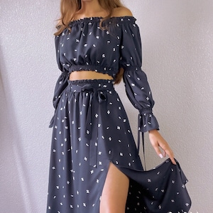May include: A black off-the-shoulder crop top with long sleeves and a white floral print. The top is paired with a matching black midi skirt with a side slit and a white floral print. The skirt has a tie at the waist.