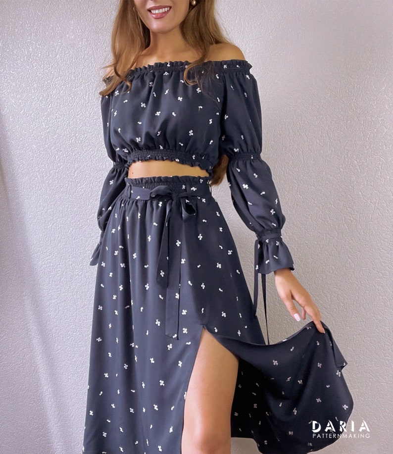 May include: A black off-the-shoulder crop top with long sleeves and a white floral print. The top is paired with a matching black midi skirt with a side slit and a white floral print. The skirt has a tie at the waist.