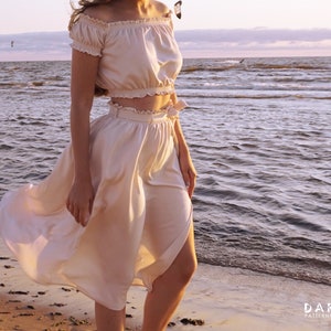May include: A woman wearing a white off-the-shoulder crop top and a matching white skirt with a wrap design. The skirt is flowing in the wind. The woman is standing on a sandy beach with the ocean in the background.
