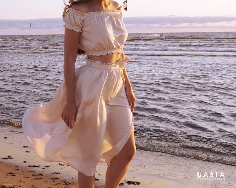 May include: A woman wearing a white off-the-shoulder crop top and a matching white skirt with a wrap design. The skirt is flowing in the wind. The woman is standing on a sandy beach with the ocean in the background.