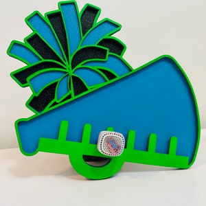 May include: A decorative cheerleader megaphone in blue, green, and black. The megaphone has a blue interior and a green outline. A small, square, bejeweled ring with a pink and blue design is attached to the megaphone.