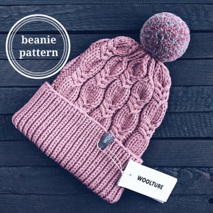 May include: A pink knitted beanie with a cable knit pattern and a fluffy pom pom on top. The beanie has a tag that says "WOOLTURE".