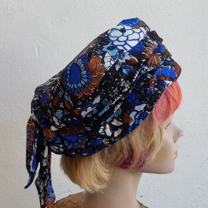 May include: A blue, brown, and white patterned turban style head scarf. The scarf is tied in a knot at the back of the head and the ends are hanging down the back. The scarf is on a mannequin with blonde hair.