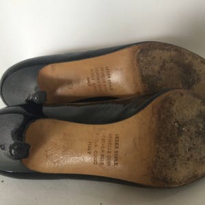 May include: A pair of black patent leather pumps with a low heel. The shoes have a leather sole and are made in Italy. The text on the sole reads "Leder Sohle Semmelle Cuir Leather Sole Suola Cuoio Italy".