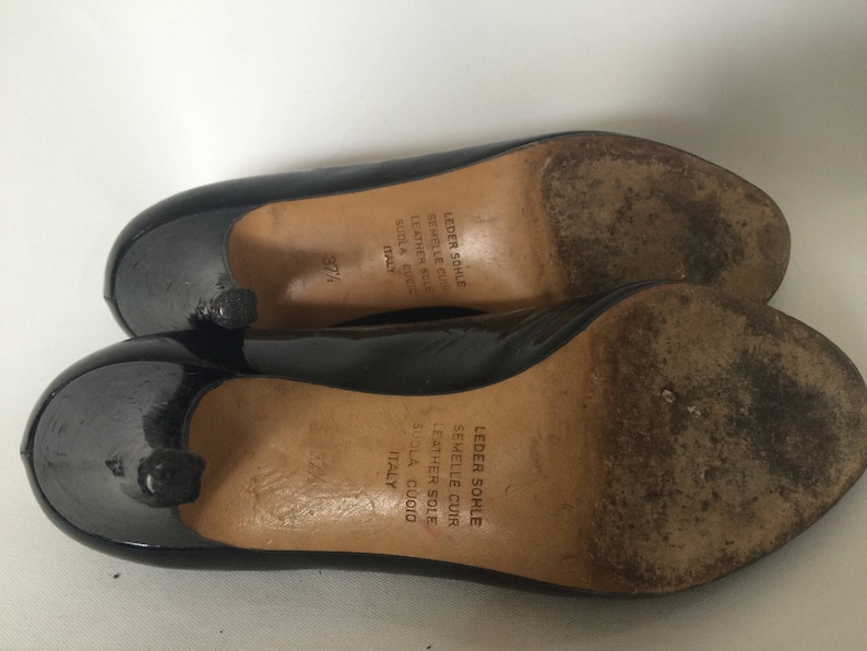 May include: A pair of black patent leather pumps with a low heel. The shoes have a leather sole and are made in Italy. The text on the sole reads "Leder Sohle Semmelle Cuir Leather Sole Suola Cuoio Italy".