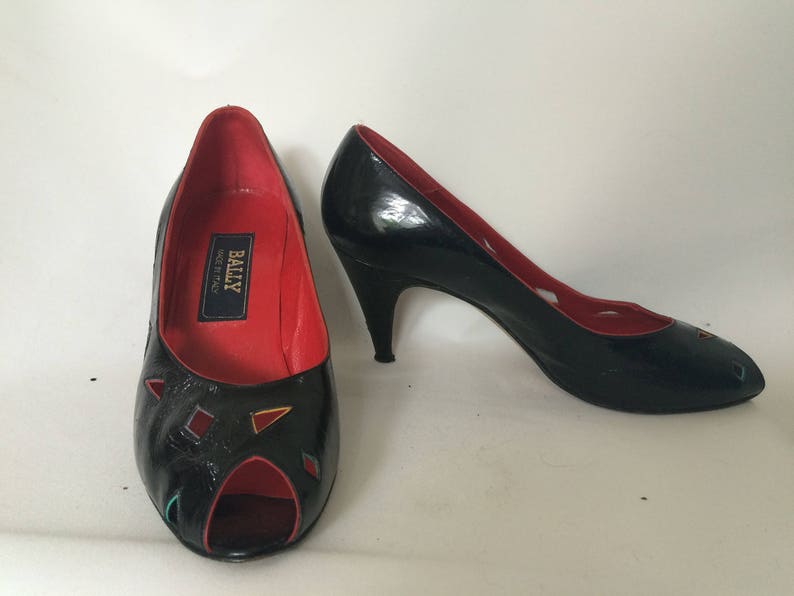 May include: A pair of black peep-toe heels with red lining and a geometric design on the toe. The heels are made by Bally, Made in Italy.