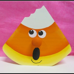 May include: A hand-painted wooden candy corn with a white top, orange middle, and yellow bottom. The candy corn has a cartoon face with big eyes, a red nose, and an open mouth.