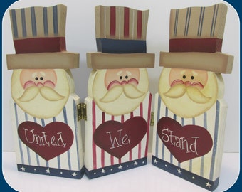 May include: Three wooden figures painted to look like Uncle Sam. Each figure has a red, white, and blue striped hat, a white beard, and a red heart with the words "United", "We", and "Stand" written on them.