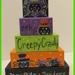 May include: A stacked Halloween decoration featuring painted wooden blocks. The blocks are orange, green, purple, and black, with spider and web designs. Text includes "Itsy Bitsy Spiders" and "Creepy Crawly."