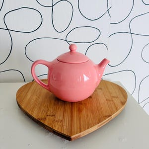 May include: A solid pink ceramic teapot with a matching lid and a curved handle, resting on a heart-shaped bamboo wood board. The teapot features a smooth, glossy finish and a rounded body with a short, pointed spout extending to the right side.