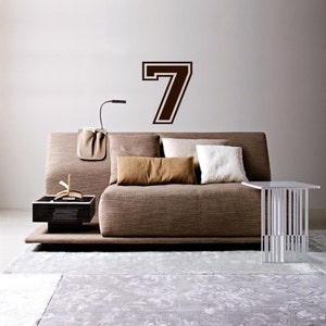 May include: A brown couch with pillows and a side table in a living room. A large brown number seven is on the wall behind the couch.