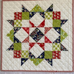 May include: A quilted star-shaped placemat with a cream background and a red and white striped border. The star design features red, green, and navy blue patterned fabric squares and triangles. The quilting pattern is a diamond grid.