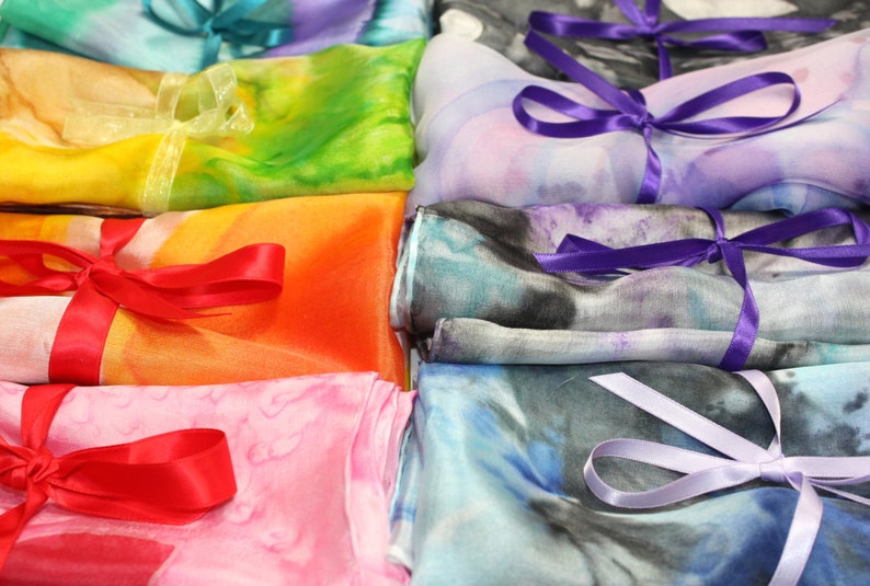 May include: A stack of colorful silk scarves with different patterns and tied with ribbons. The scarves are arranged in a rainbow order, with red, orange, yellow, green, blue, and purple.