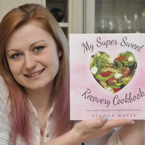 May include: A woman holds a pink cookbook titled "My Super Sweet Recovery Cookbook" with a heart-shaped window showing a salad inside. The cookbook cover also includes the text "Recipes designed to help people with eating disorders in their recovery" and "NICOLA DAVIS".