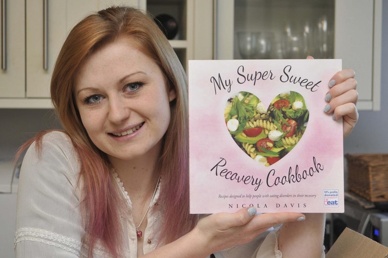 May include: A woman holds a pink cookbook titled "My Super Sweet Recovery Cookbook" with a heart-shaped window showing a salad inside. The cookbook cover also includes the text "Recipes designed to help people with eating disorders in their recovery" and "NICOLA DAVIS".