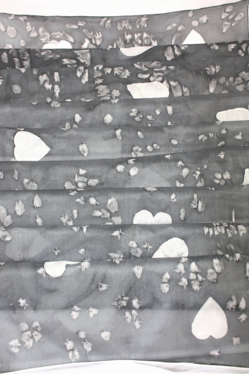 Peut inclure: Une &eacute;charpe en soie grise avec un motif de c&oelig;urs blancs. L'&eacute;charpe est pli&eacute;e en plusieurs couches, cr&eacute;ant un aspect textur&eacute;.
