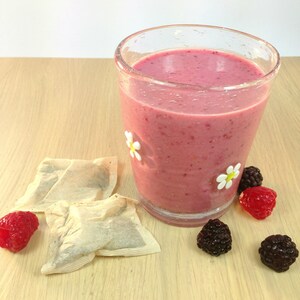 May include: A glass of pink smoothie with a white daisy design on the side. The smoothie is surrounded by raspberries, blackberries, and tea bags on a wooden surface.