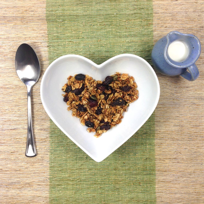 May include: A white heart-shaped bowl filled with granola, cranberries, and nuts. The bowl is on a green and brown woven placemat. A small blue pitcher with milk is in the background.