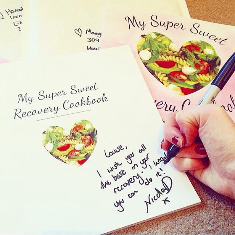 May include: A white cookbook with a heart-shaped cutout revealing a salad. The title of the book is "My Super Sweet Recovery Cookbook". The book is signed with a message that reads "Louise, I wish you all the best in your recovery. I know you can do it! Nichola".