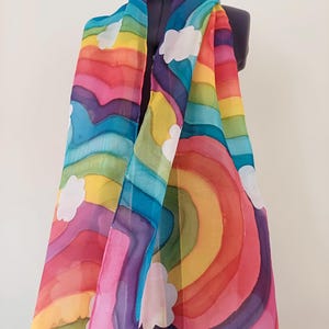 May include: A colorful silk scarf with a rainbow and cloud design. The scarf is a vibrant mix of pink, red, orange, yellow, green, blue, and purple.
