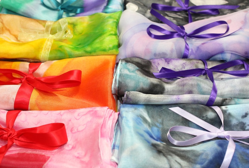 May include: A stack of colorful silk scarves with different patterns and tied with ribbons. The scarves are arranged in a rainbow order, with red, orange, yellow, green, blue, and purple.