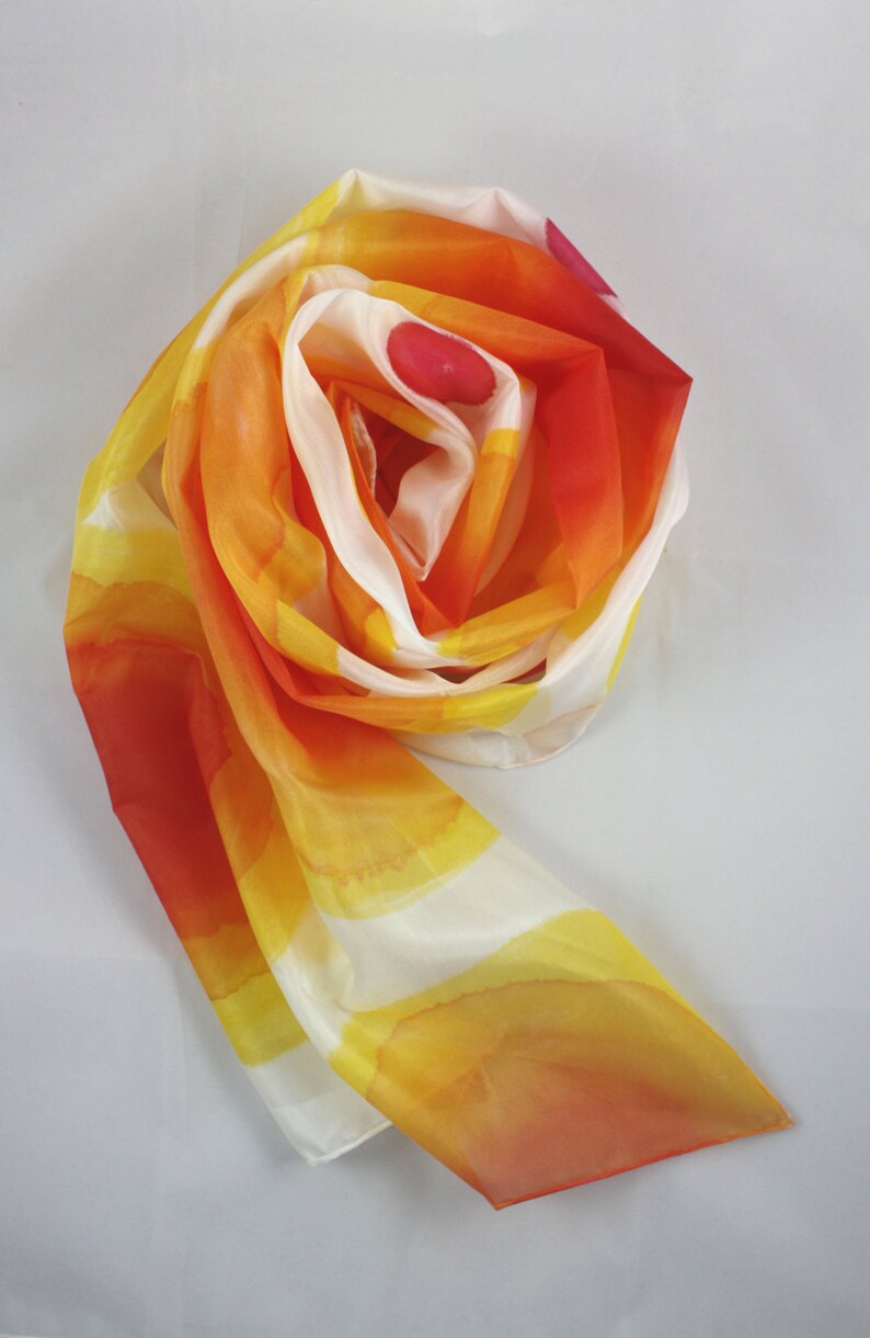 May include: A white silk scarf with orange, yellow, and red watercolor-like designs. The scarf is folded and tied into a loose knot.