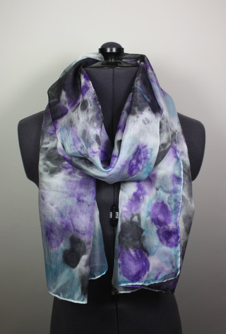May include: A light blue silk scarf with a hand-painted design of purple and black watercolor-like shapes.